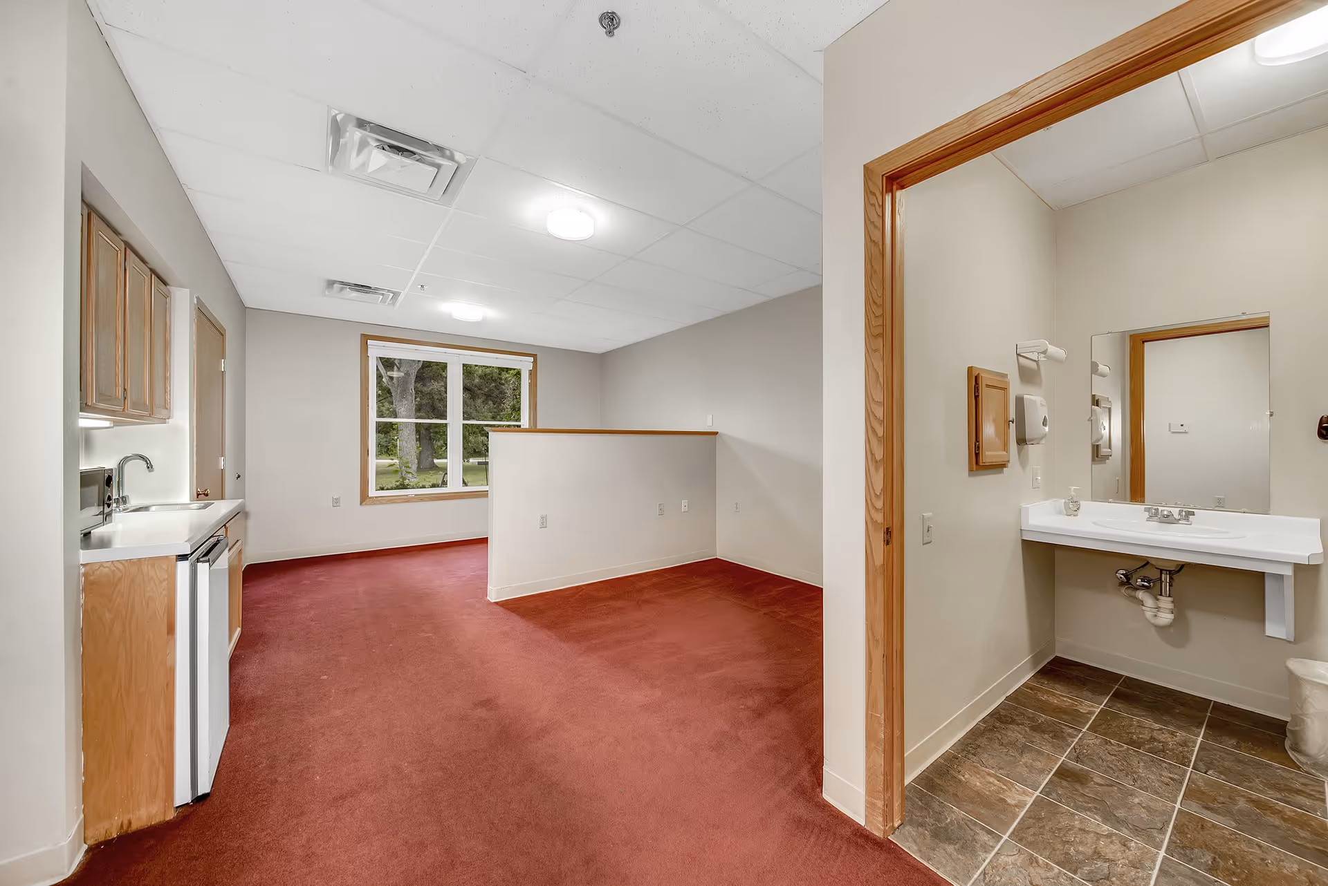 Interior view of a senior living facility room with a small kitchenette on the left, featuring a sink, mini fridge, and cabinets. The room has red carpet flooring and a large window with a view of trees outside. To the right, there is an open doorway leading to a bathroom area with a wall-mounted sink, mirror, and tiled floor.