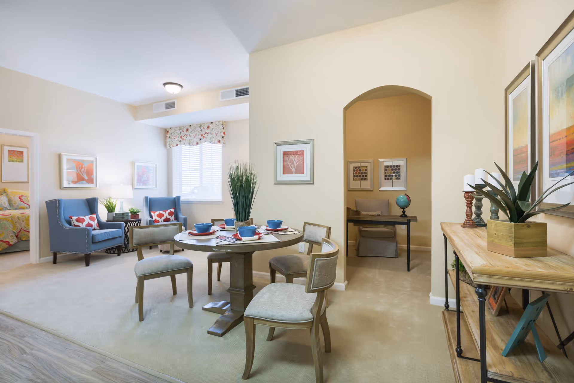 A bright and cozy living and dining area in a senior living facility. The room features a round dining table set with blue bowls and red napkins, surrounded by four cushioned chairs. To the left, there are two blue armchairs with red and white patterned pillows next to a window with floral valance. The walls are decorated with framed artwork, and a wooden console table with plants and candles is on the right. An arched doorway leads to a small office nook with a chair, desk, and globe.