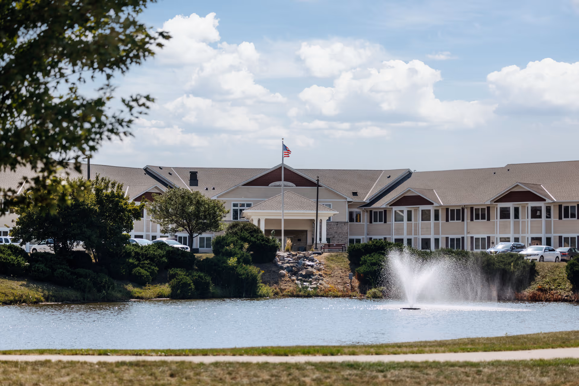 Exterior front view of a multi-story assisted living building with an American flag and a pond with a fountain in the foreground.