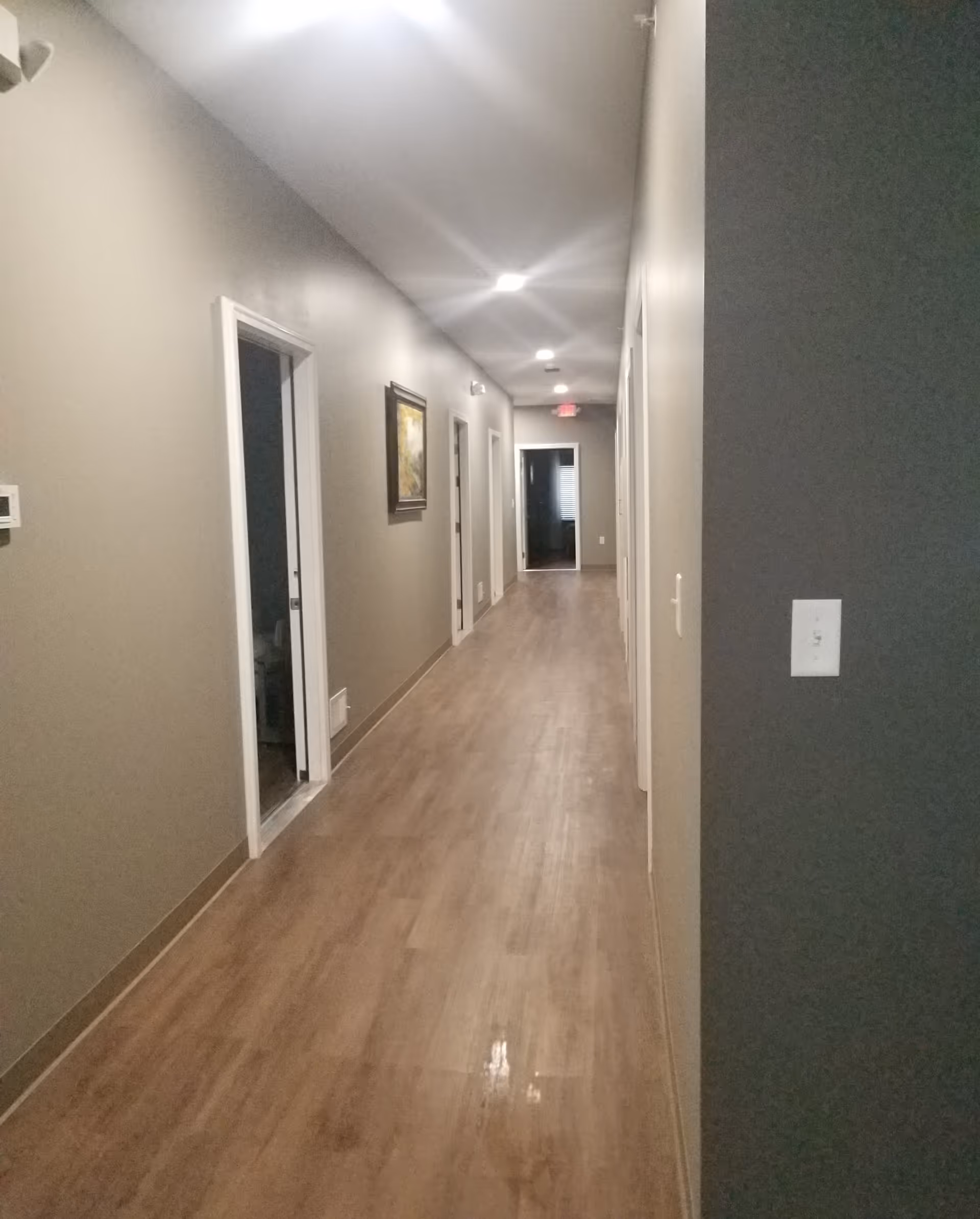 Long interior hallway with wood-look flooring, recessed ceiling lights, and several doorways along the walls.