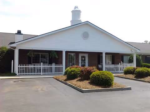 Front exterior view of a single-story brick building with a white porch, white picket fence, and a small landscaped area with bushes in front. The building has a cupola on the roof and a paved driveway or parking area in front.
