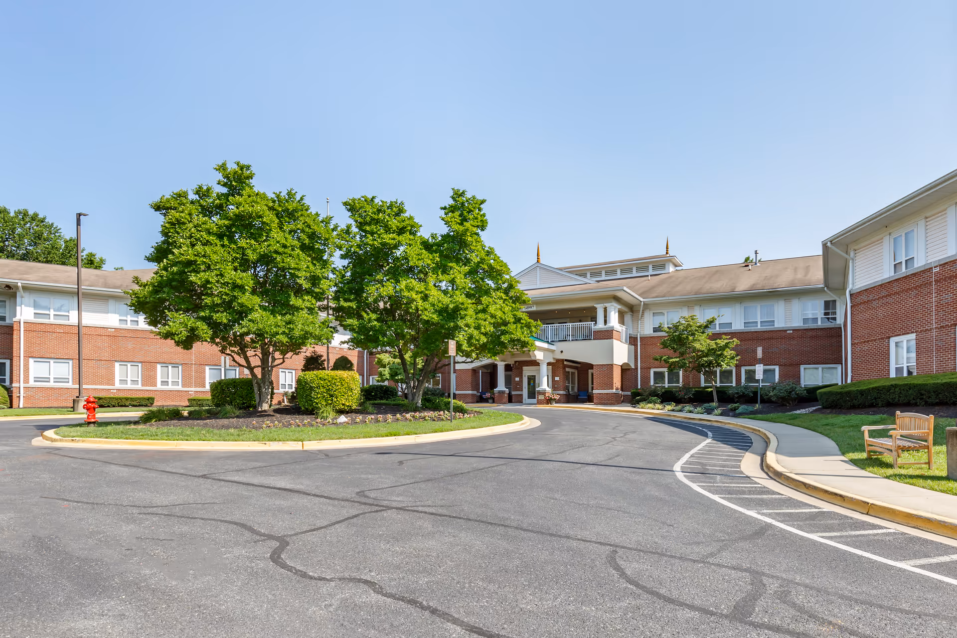 Exterior view of a senior living facility with a circular driveway, green trees, bushes, and a bench on the lawn. The building has red brick walls and white trim under a clear blue sky.