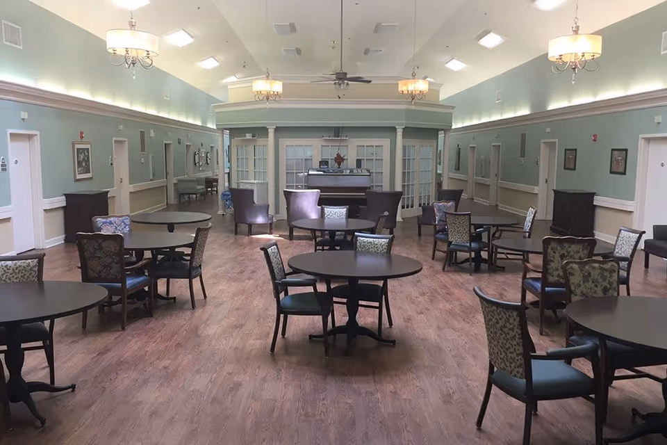 Spacious senior living common room with round tables and chairs, high ceiling, soft green walls, and a central seating area.
