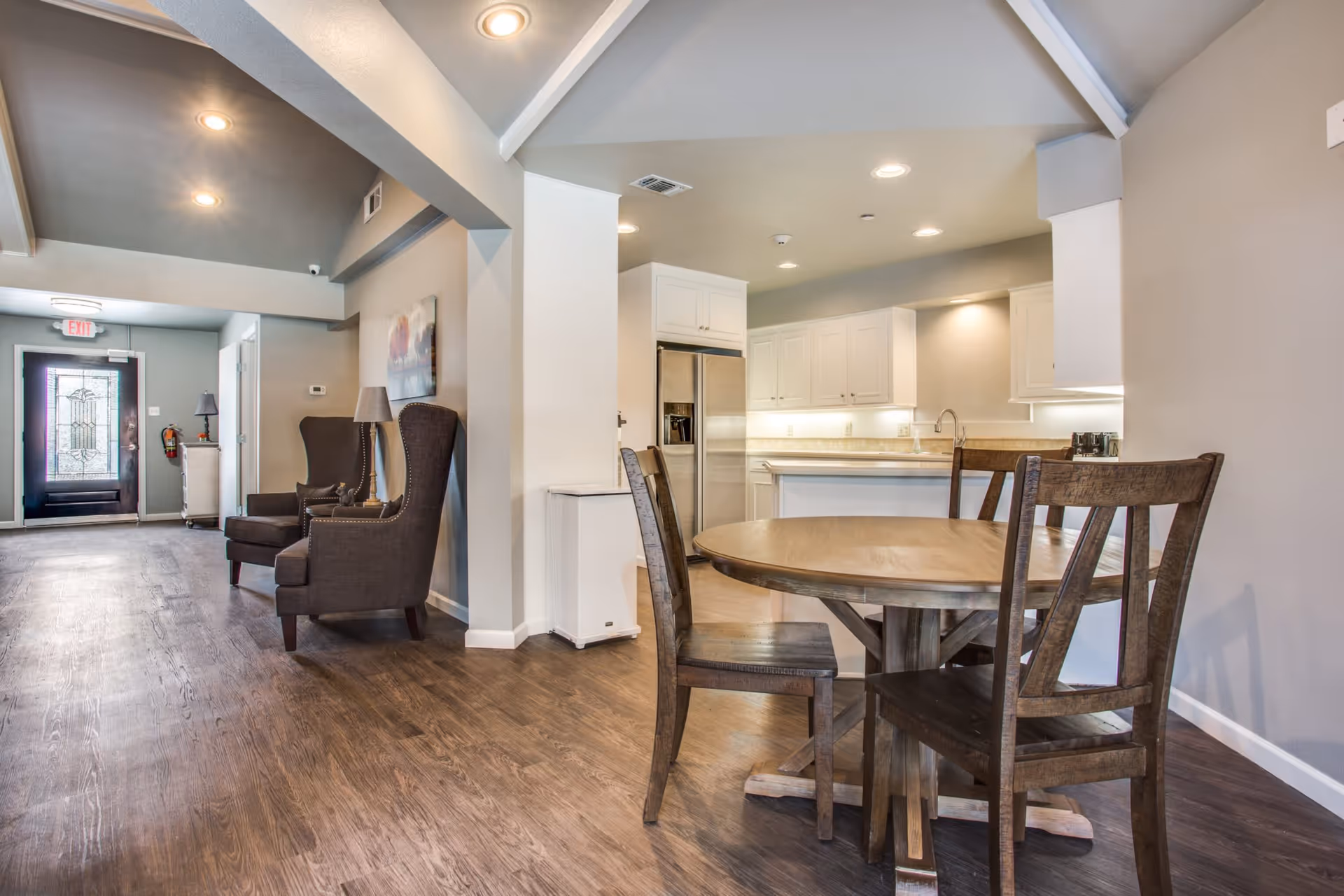 Interior view of a senior living facility showing a dining area with a round wooden table and four chairs, adjacent to a kitchen with white cabinets and stainless steel refrigerator. There are two dark upholstered armchairs with a lamp between them along the hallway leading to a door with a decorative glass panel.