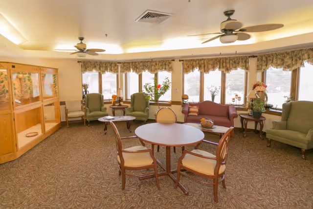 A spacious living room area with patterned carpet, a round table with four chairs in the center, two green armchairs, a maroon sofa, side tables with plants and decorative items, large windows with floral valances, and ceiling fans.