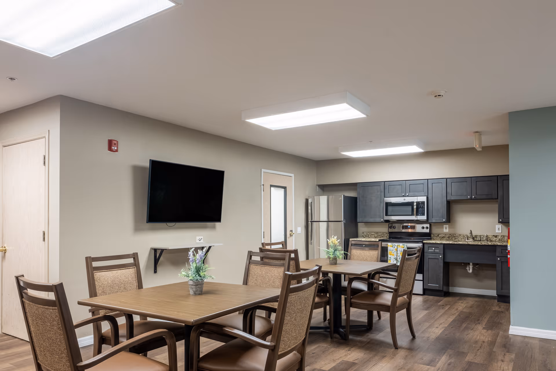 Interior view of a senior living facility common area with a kitchen and dining space. The room features two wooden tables with chairs around them, a wall-mounted flat-screen TV, and a kitchen area with dark cabinets, a stainless steel refrigerator, microwave, and stove. The floor is wood, and the walls are painted in neutral tones with bright ceiling lights.