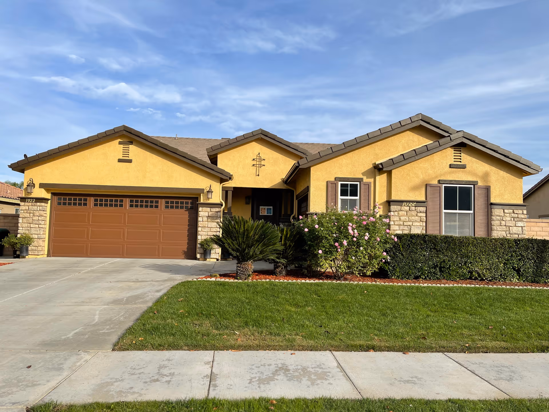Front exterior view of a single-story yellow house with a brown garage door, stone accents, and a well-maintained lawn with bushes and flowers under a partly cloudy sky.