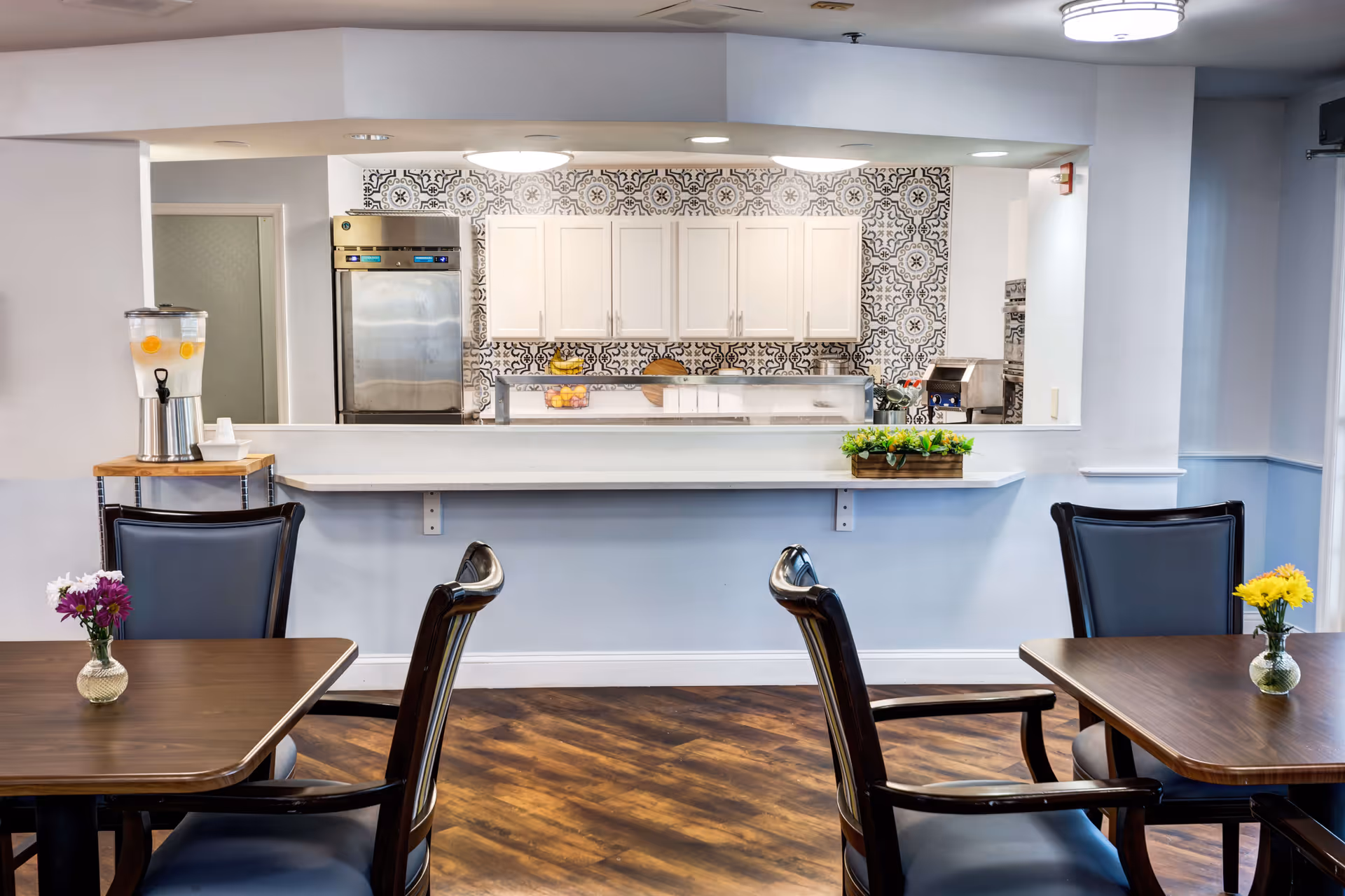 Dining area with wooden tables and chairs, each table decorated with a small vase of flowers. In the background, a kitchen area with white cabinets, a stainless steel refrigerator, and a patterned tile backsplash is visible through a serving window. A water dispenser with lemon slices is placed on a small table near the serving window.