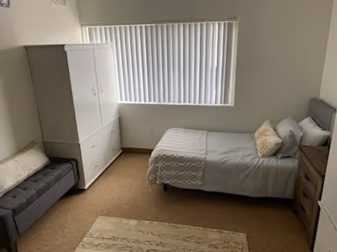 A simple bedroom with a single bed made with gray and white bedding, a wooden nightstand with a lamp, a white wardrobe, a gray cushioned bench, and a window with vertical blinds letting in natural light.