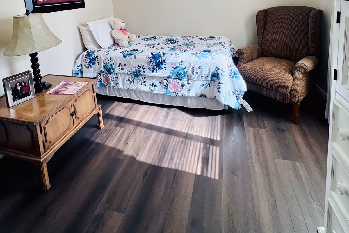 A cozy bedroom with a single bed covered in a floral bedspread and a teddy bear placed on the pillow. Next to the bed is a wooden nightstand with a lamp, a framed photo, and a magazine. There is a comfortable brown armchair in the corner and sunlight casting shadows on the wooden floor.