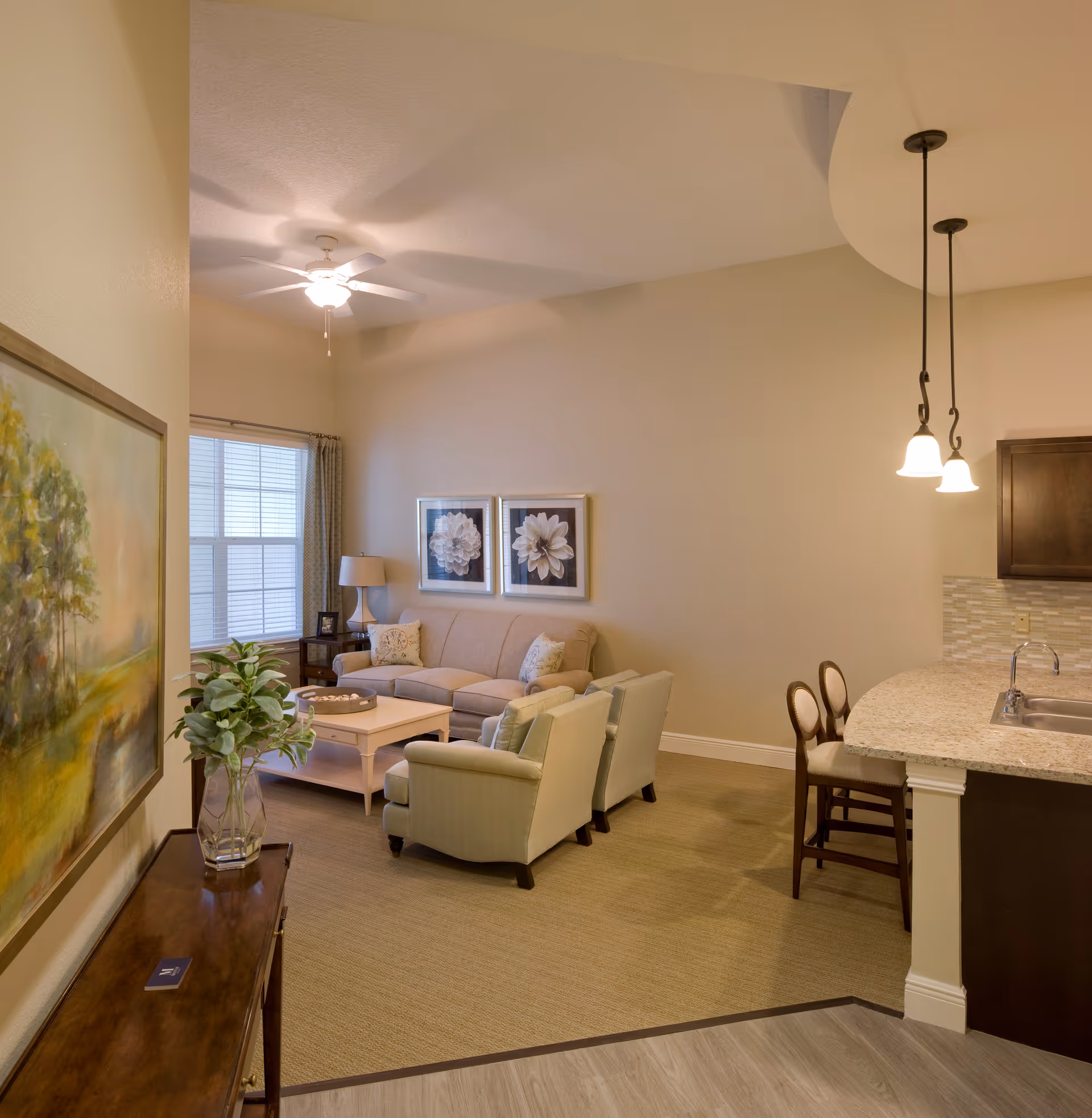 A cozy living room area with a beige sofa, two light green armchairs, a white coffee table, and a side table with a lamp. The room has a ceiling fan, two framed floral pictures on the wall, and a window with curtains. Adjacent to the living area is a kitchen counter with two chairs and pendant lights hanging above.