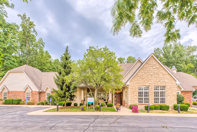 Exterior view of a single-story assisted living facility building with a mix of brick and stone facade, surrounded by trees and greenery under a cloudy sky. There is a paved driveway and walkway leading to the entrance, with some landscaping including bushes and flowers.