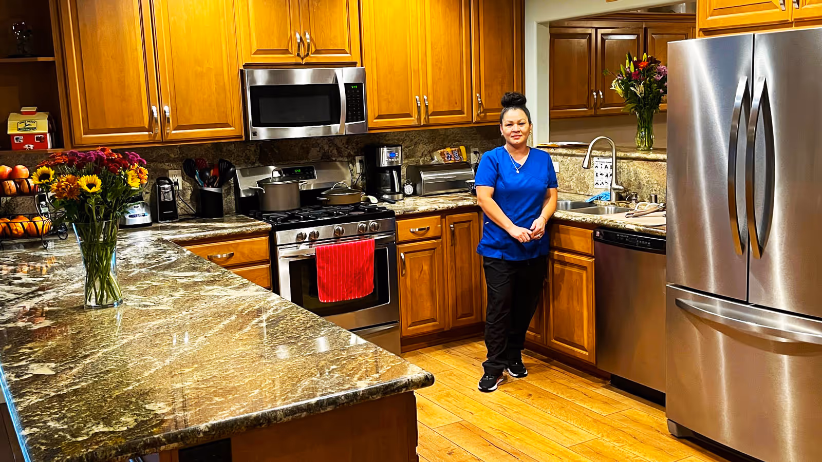 A woman in blue scrubs standing in a spacious kitchen with wooden cabinets, granite countertops, stainless steel appliances including a refrigerator, microwave, stove, and dishwasher. There is a vase with flowers on the counter and various kitchen items around.