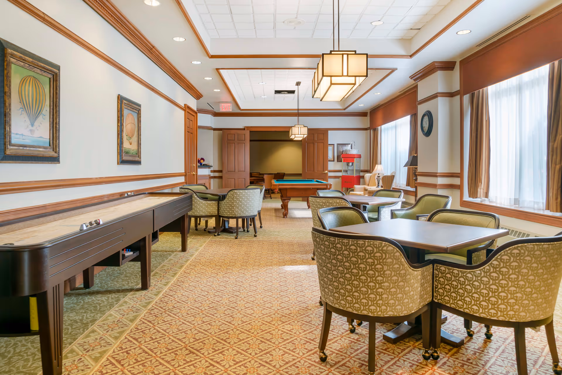 A well-lit recreational room with patterned carpet, several tables surrounded by upholstered chairs, a shuffleboard table along the left wall, and a pool table in the background. The room has large windows with curtains, framed hot air balloon artwork on the walls, and hanging rectangular light fixtures from the ceiling.