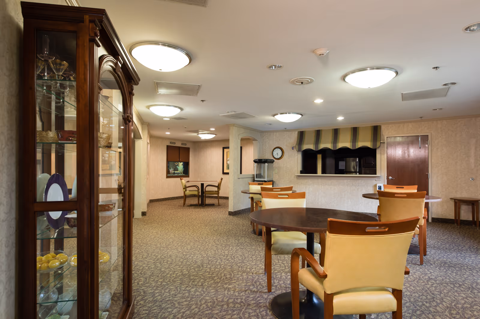 A well-lit communal dining area with round wooden tables, upholstered chairs, a glass display cabinet, and a serving window.