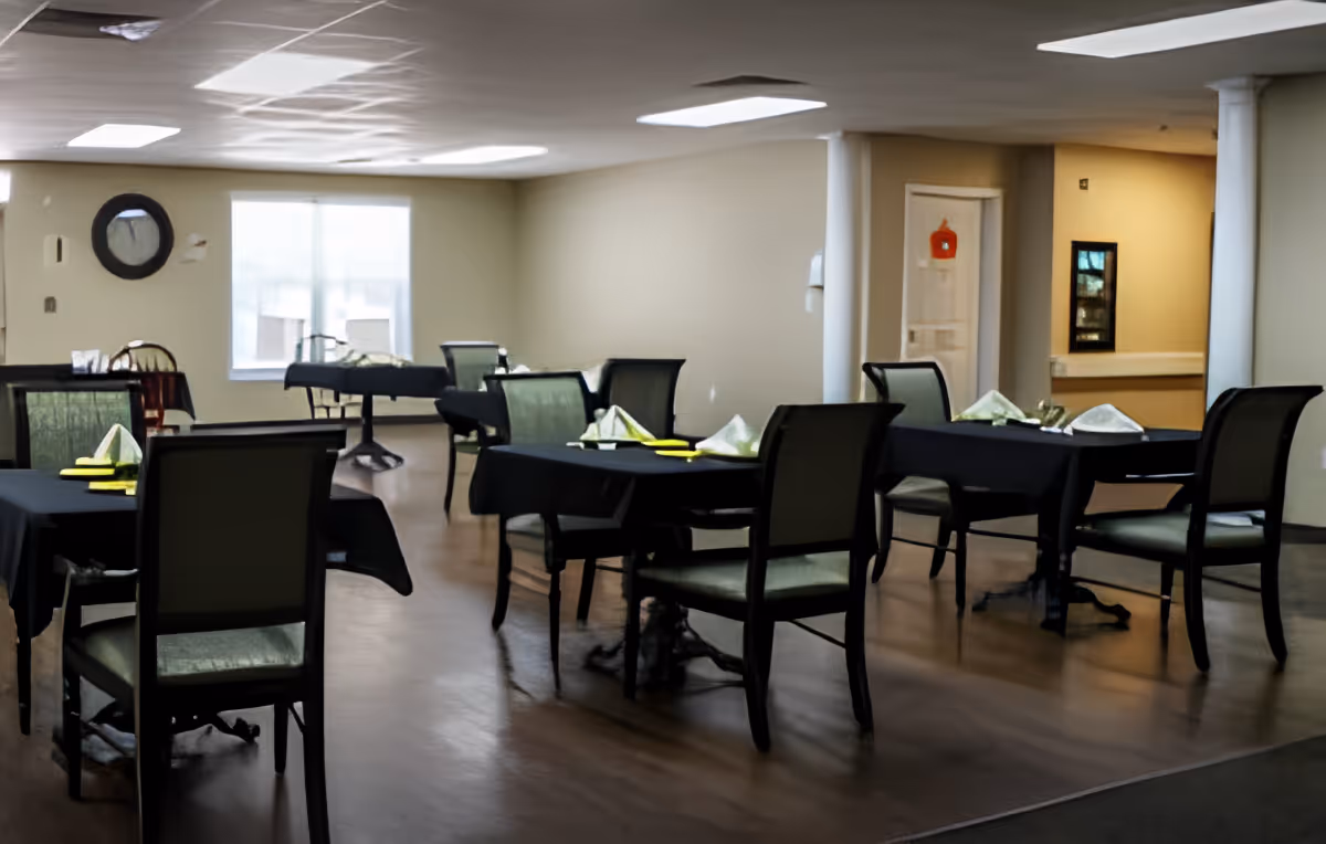 Dining room with several tables covered with black tablecloths, each set with folded napkins and utensils. Chairs with green upholstery surround the tables. The room has beige walls, a window with blinds, and white columns.