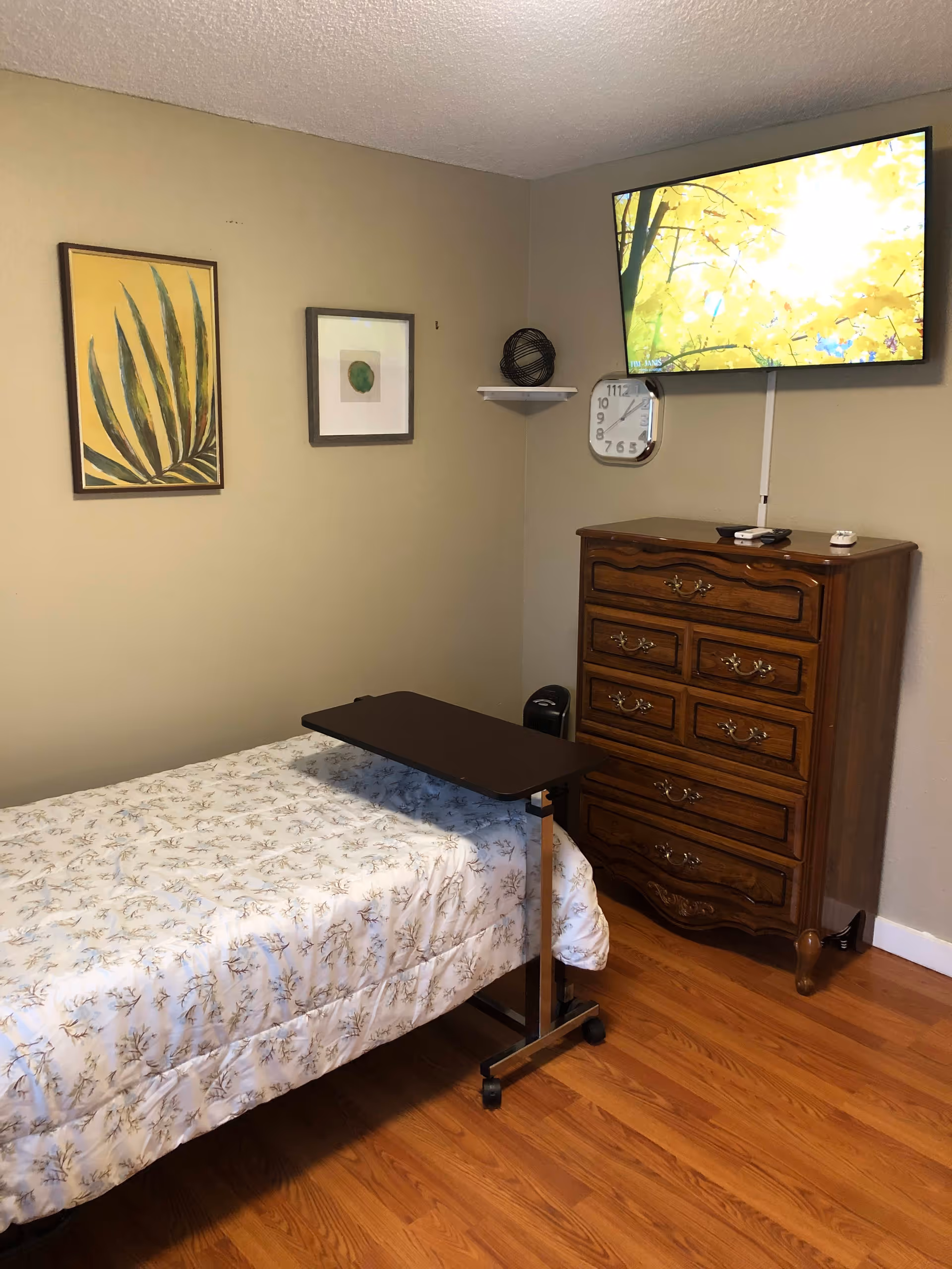 A bedroom with a single bed covered in a white floral bedspread, a small adjustable overbed table on wheels, a wooden dresser with multiple drawers, a wall-mounted flat-screen TV displaying a bright image of yellow leaves, a wall clock, and two framed pictures on the beige wall.
