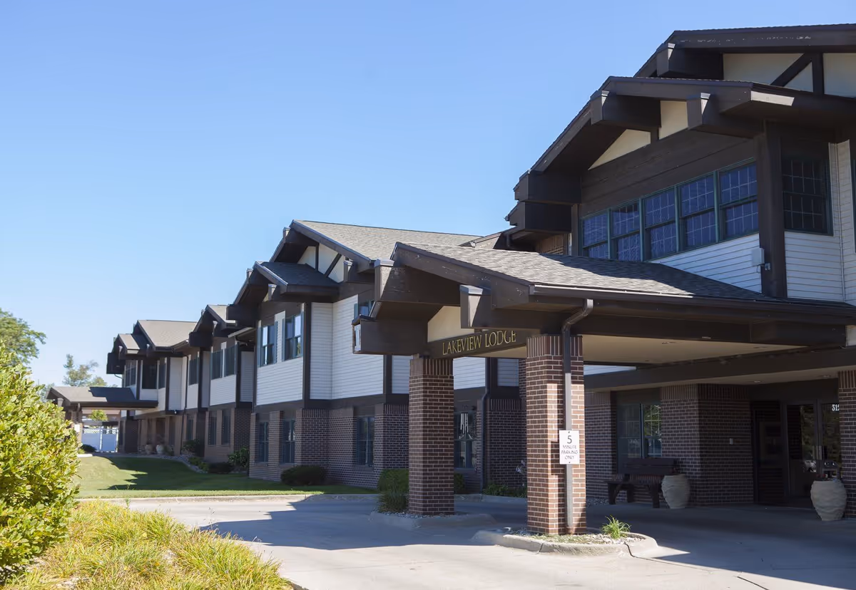 Exterior view of Lakeview Lodge Assisted Living at Friendship Village, showing a two-story building with a covered entrance supported by brick columns under a clear blue sky.