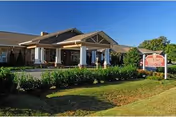 Front exterior of the Webb House Retirement Center showing the brick building's covered entrance, landscaped bushes, lawn, and a roadside sign under a clear blue sky.