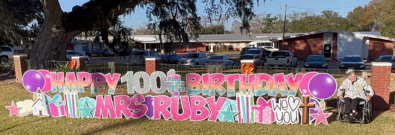 Outdoor scene with a large colorful sign on the grass that reads 'Happy 100th Birthday Mrs Ruby We Love You' decorated with balloons, stars, gifts, and candles. An elderly woman in a wheelchair is positioned to the right of the sign. In the background, there are parked cars and a single-story building under a clear sky.