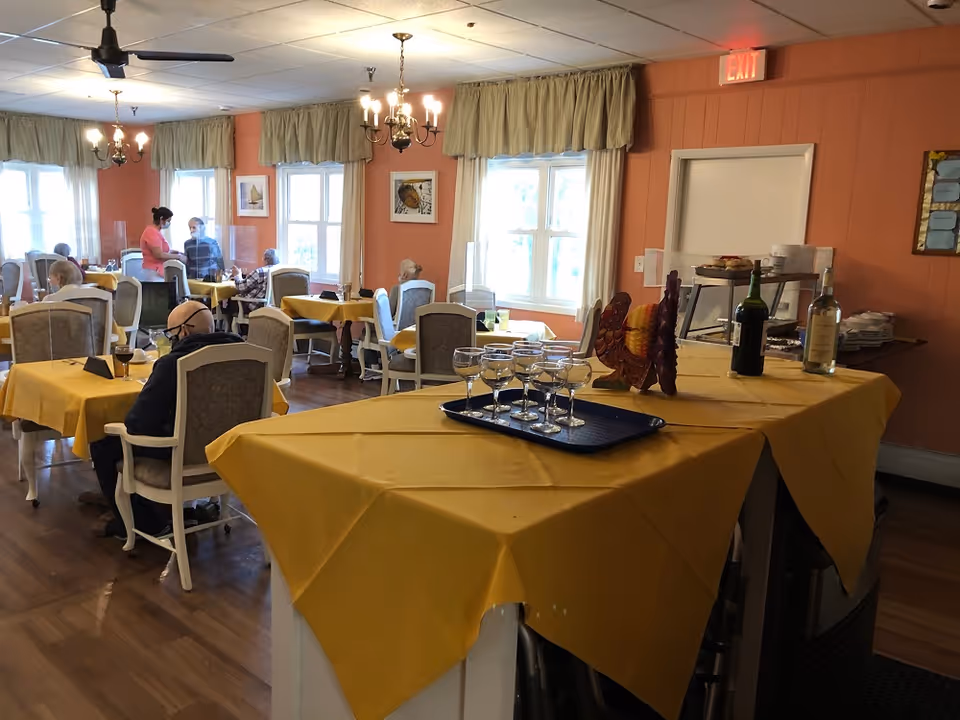 Dining room with yellow-tablecloth covered tables, several seated senior residents and staff, and a serving area with glasses and bottles in the foreground.