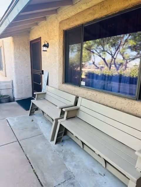 Outdoor seating area with two beige benches placed against a textured beige wall of a building. A door with a screen and a welcome sign is visible, along with a window reflecting trees and sky. The area is paved with concrete.