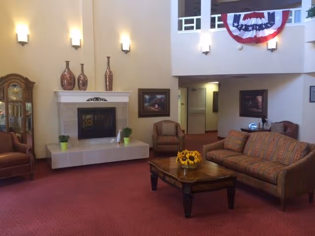 Spacious retirement community lounge with sofa and armchairs around a coffee table, a fireplace, and an upper balcony draped with patriotic bunting.