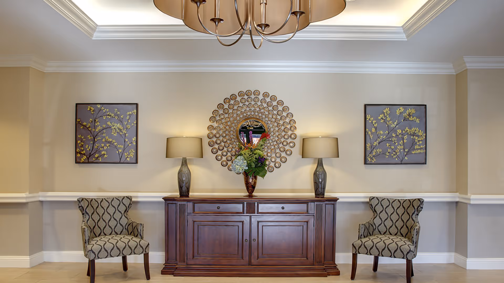 Symmetrical seating area with two patterned chairs flanking a wooden console topped with lamps, a decorative round mirror, and a floral arrangement.