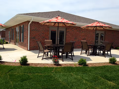 Outdoor patio area with round tables, chairs, and red patterned umbrellas next to a single-story brick building under a partly cloudy sky. The patio is surrounded by green grass and small shrubs.