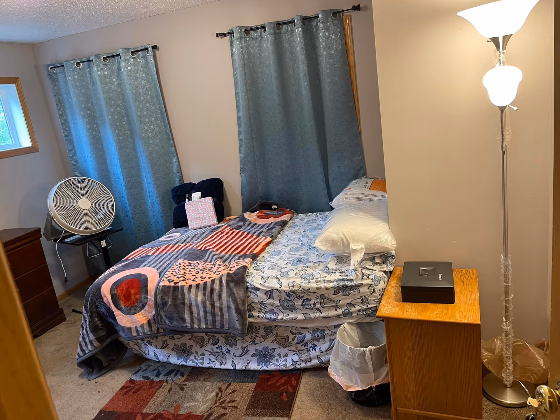 A small bedroom with a bed covered in floral sheets and a colorful blanket. There are two pillows on the bed, a wooden nightstand with a black box on it, a standing lamp, a small trash can, a fan on a stand, and blue curtains covering the windows.