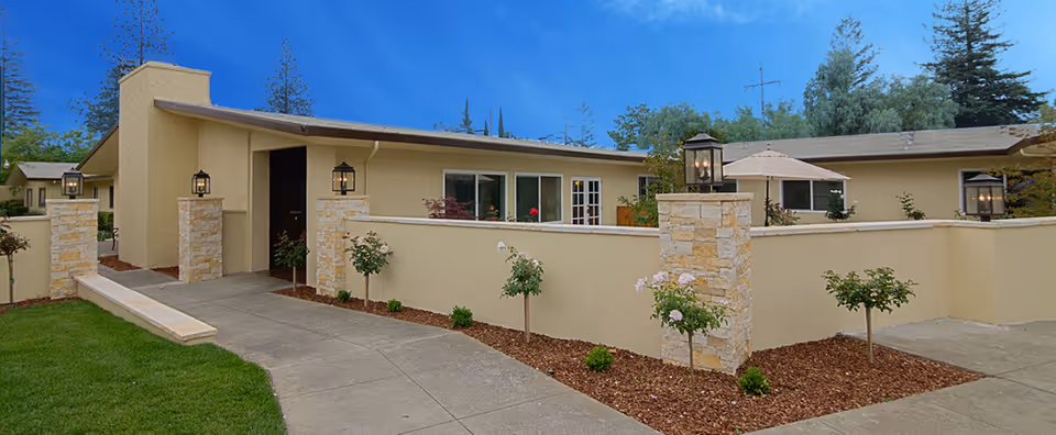 Single-story beige senior living facility front with a low courtyard wall, stone pillars with lanterns, landscaping and a concrete walkway.