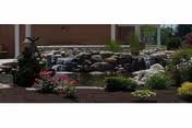 Outdoor garden area with a small waterfall flowing over rocks into a pond, surrounded by various plants and flowers, with a brick building partially visible in the background.