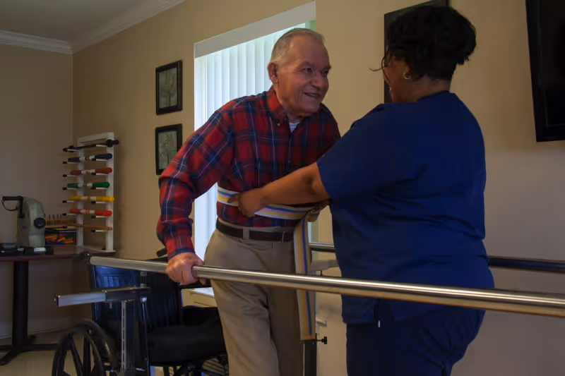 An elderly man wearing a red plaid shirt and beige pants is using parallel bars for support while a caregiver in blue scrubs assists him by holding a gait belt around his waist. A wheelchair and exercise equipment are visible in the background inside a room with beige walls.