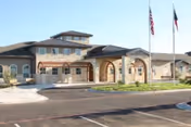 Exterior view of a single-story senior living facility building with a covered entrance, arched stone columns, and two flagpoles displaying the American and Texas flags in front. The building has a combination of stone and siding exterior with multiple windows and a clear blue sky above.