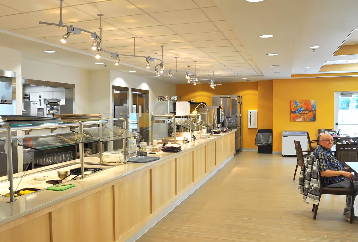 Interior view of a senior living facility dining area with a long serving counter featuring glass sneeze guards and trays. A man is seated at a table on the right side near a window, with yellow walls and a colorful painting in the background.