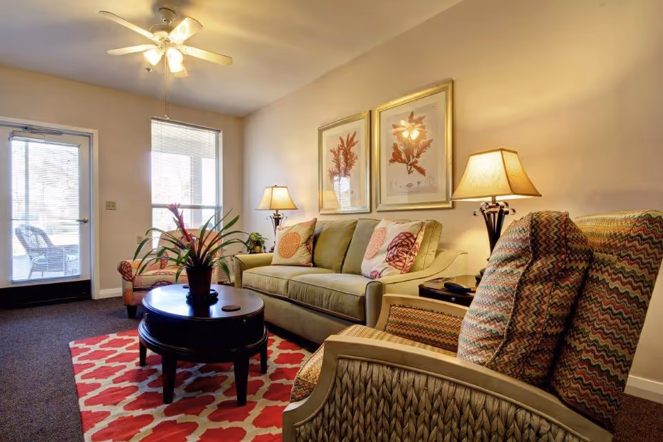 Bright living room with a sofa, patterned armchairs, coffee table, lamps, framed art, and a red patterned rug.