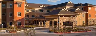 Exterior view of Discovery Village Stone Oak, a multi-story senior living facility with a combination of stone and stucco facade, multiple windows, and a covered entrance. The building is surrounded by landscaped grounds and a driveway.