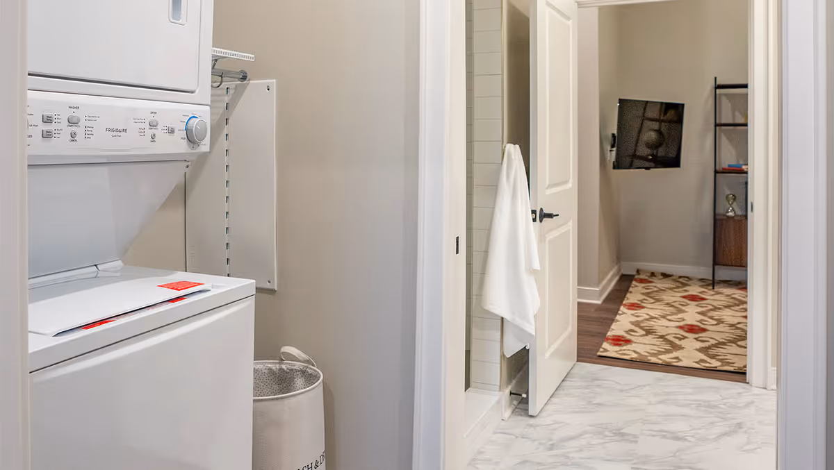 A laundry area with a stacked washer and dryer unit on the left, a laundry basket below, and a hallway leading to a room with a wall-mounted TV and a patterned rug. A white towel hangs on a door near the laundry area.