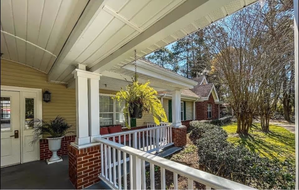 A covered porch area of a residential building with white railings and brick pillars. There is a hanging fern plant, red cushioned seating, and potted plants near the entrance door. The porch overlooks a garden area with bushes and trees under a clear blue sky.