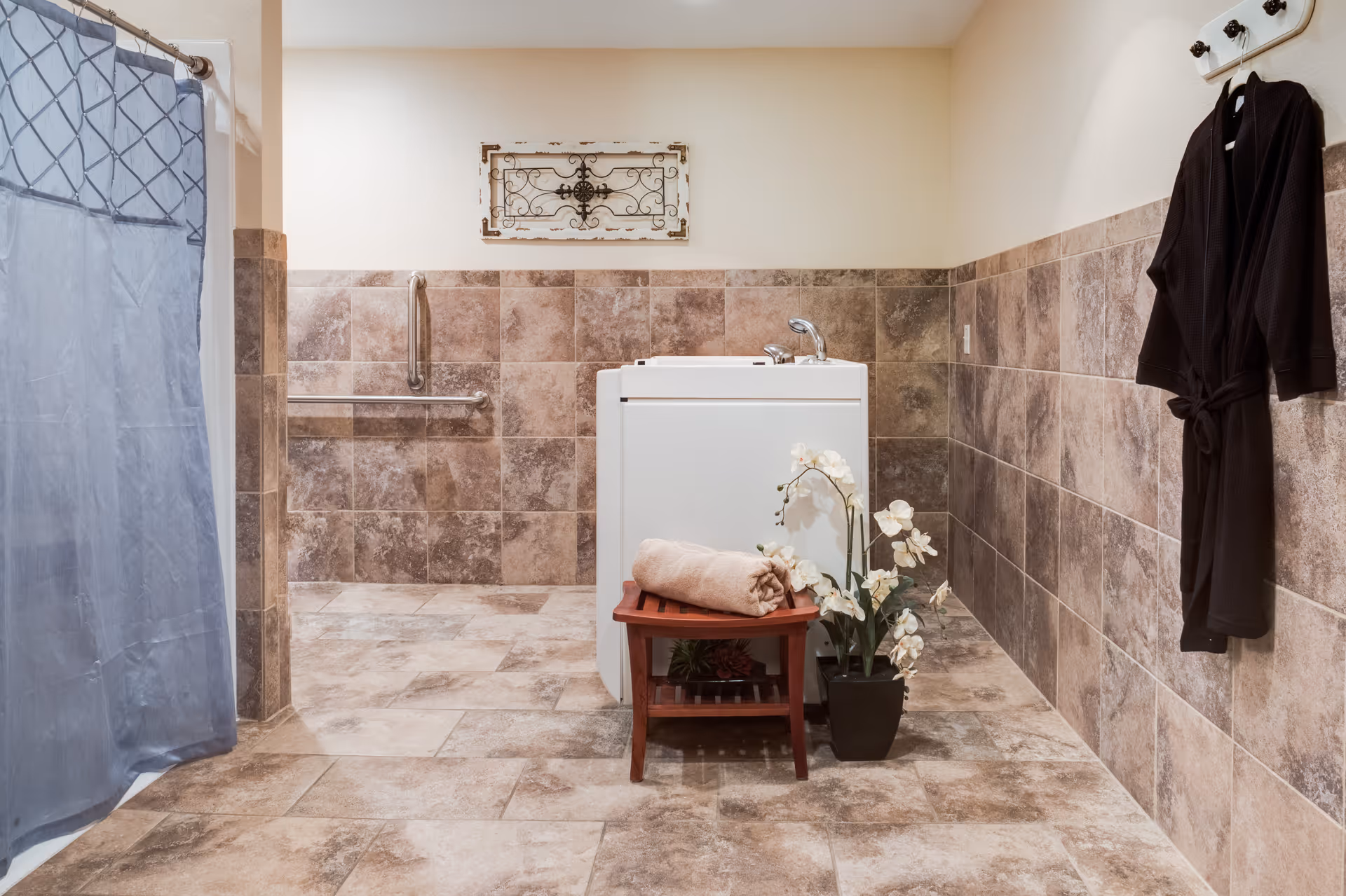 A bathroom with beige and brown tiled walls and floor. There is a white walk-in bathtub in the center with a small wooden bench in front of it holding a rolled beige towel and a potted white orchid plant. A dark robe hangs on hooks on the right wall, and a blue shower curtain is partially visible on the left. There are metal grab bars on the back wall and a decorative metal wall hanging above the bathtub.