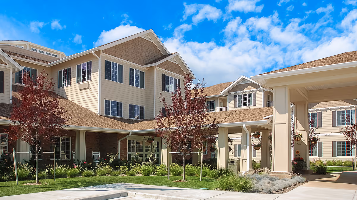 Exterior view of a senior living facility building with beige siding, multiple windows with dark shutters, and a covered entrance. The foreground features a landscaped area with small trees, green grass, and flowering plants under a bright blue sky with scattered clouds.