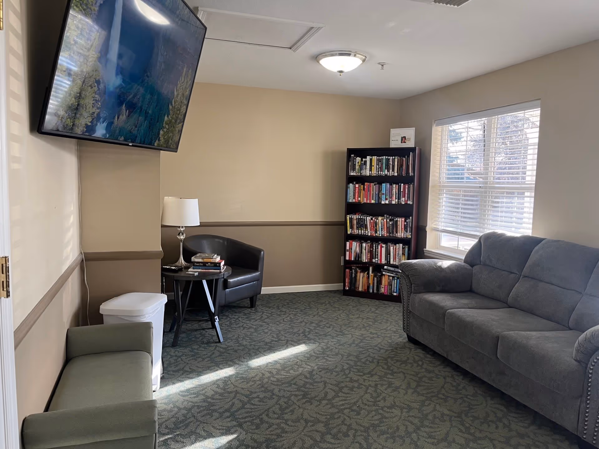 A cozy living room area with a gray couch, a black armchair, a small round table with a lamp and books, a bookshelf filled with books, a large window with blinds, and a wall-mounted flat screen TV.