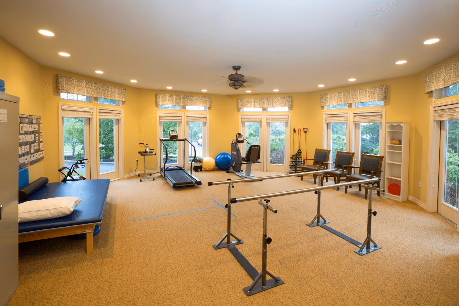 A well-lit physical therapy or exercise room with yellow walls and multiple windows. The room contains exercise equipment including a treadmill, stationary bike, exercise balls, parallel bars for walking support, a padded therapy table with a pillow, and several chairs arranged along the wall. There is also a ceiling fan and a white shelving unit with some items on it.