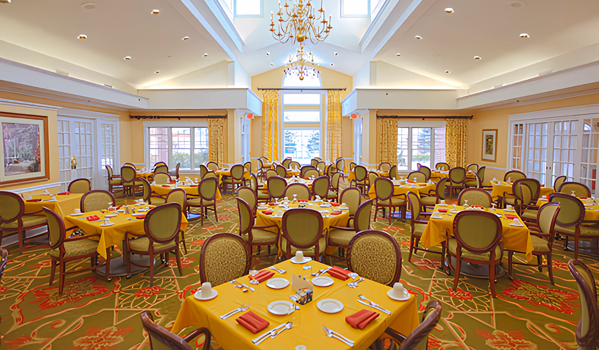 Large bright dining room with many round tables covered in yellow tablecloths set with plates and red napkins, surrounded by upholstered chairs under skylights and a chandelier.