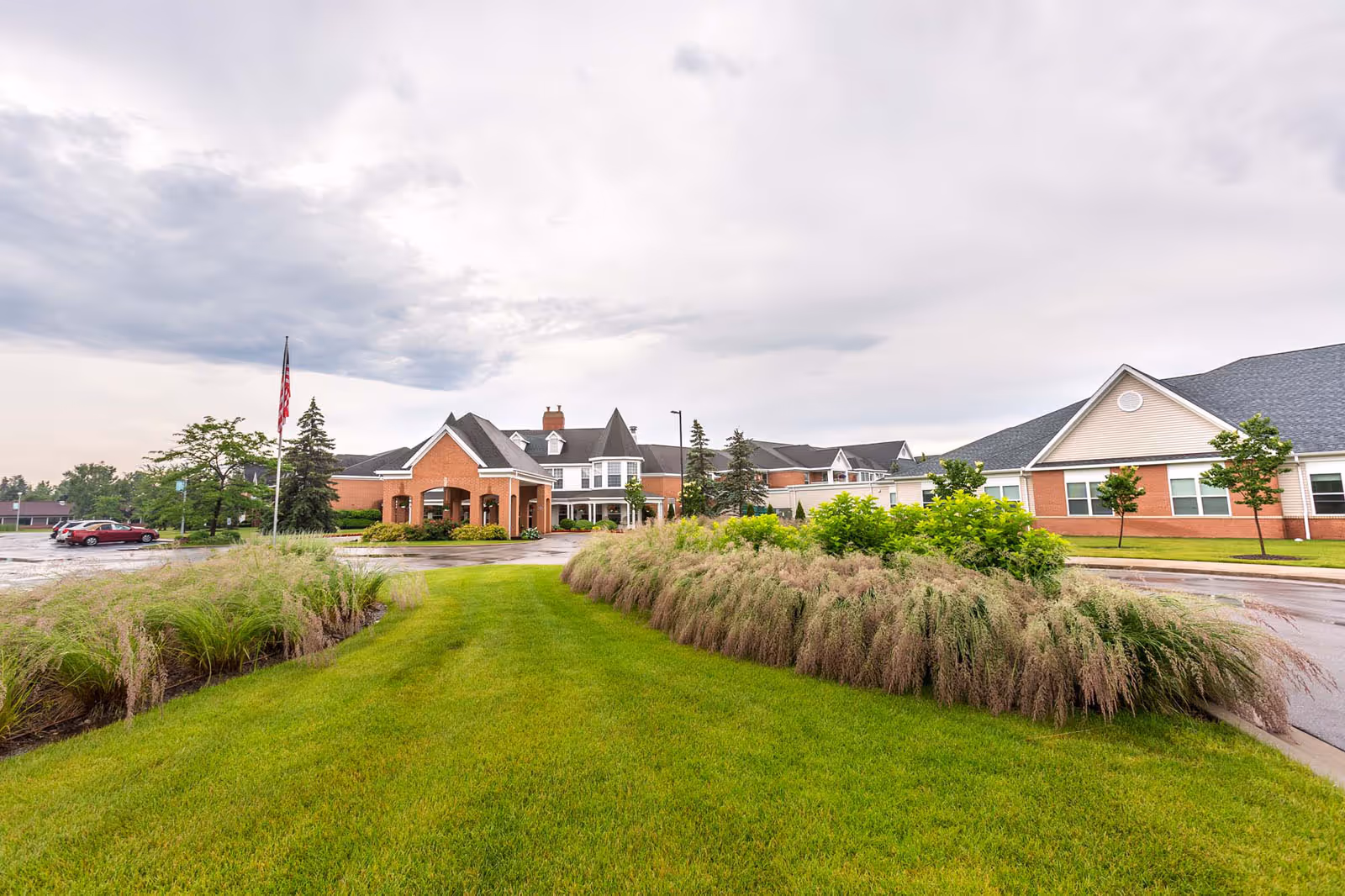 Exterior view of Independence Village Of Midland showing a large brick building with a covered entrance, surrounded by well-maintained green lawns, ornamental grasses, small trees, and a parking lot with a few cars. The sky is cloudy.