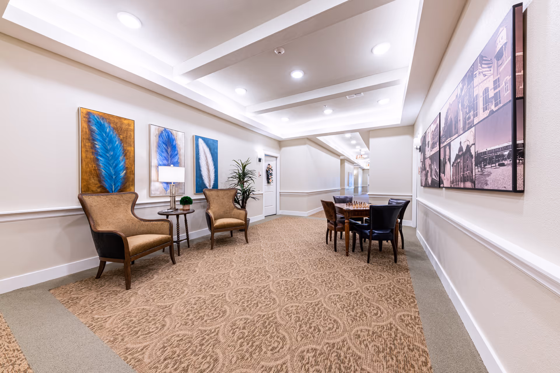 A well-lit hallway area in a senior living facility with two upholstered armchairs and a small round table with a lamp and plant on the left side. The walls are decorated with three framed feather artworks in blue and white tones. On the right side, there is a table with four chairs arranged around it, featuring a chessboard. The hallway has patterned carpet flooring and white walls with molding, and recessed ceiling lights provide illumination.