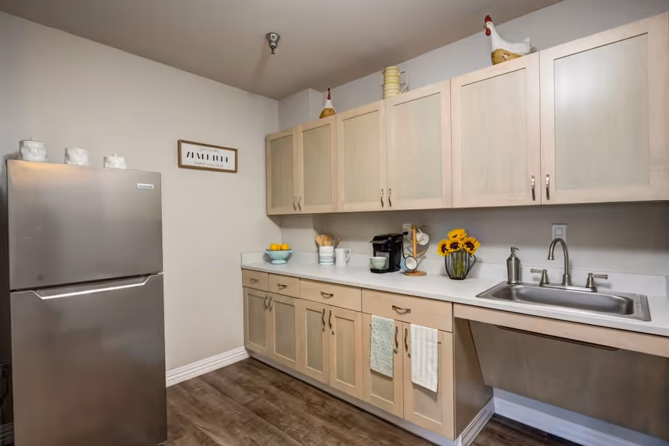 A kitchen area with light wood cabinets, a stainless steel refrigerator, a countertop with a coffee maker, mugs, a bowl of lemons, a vase with sunflowers, and a sink with a faucet. The floor has a wood finish, and there are decorative items on top of the cabinets including ceramic chickens.