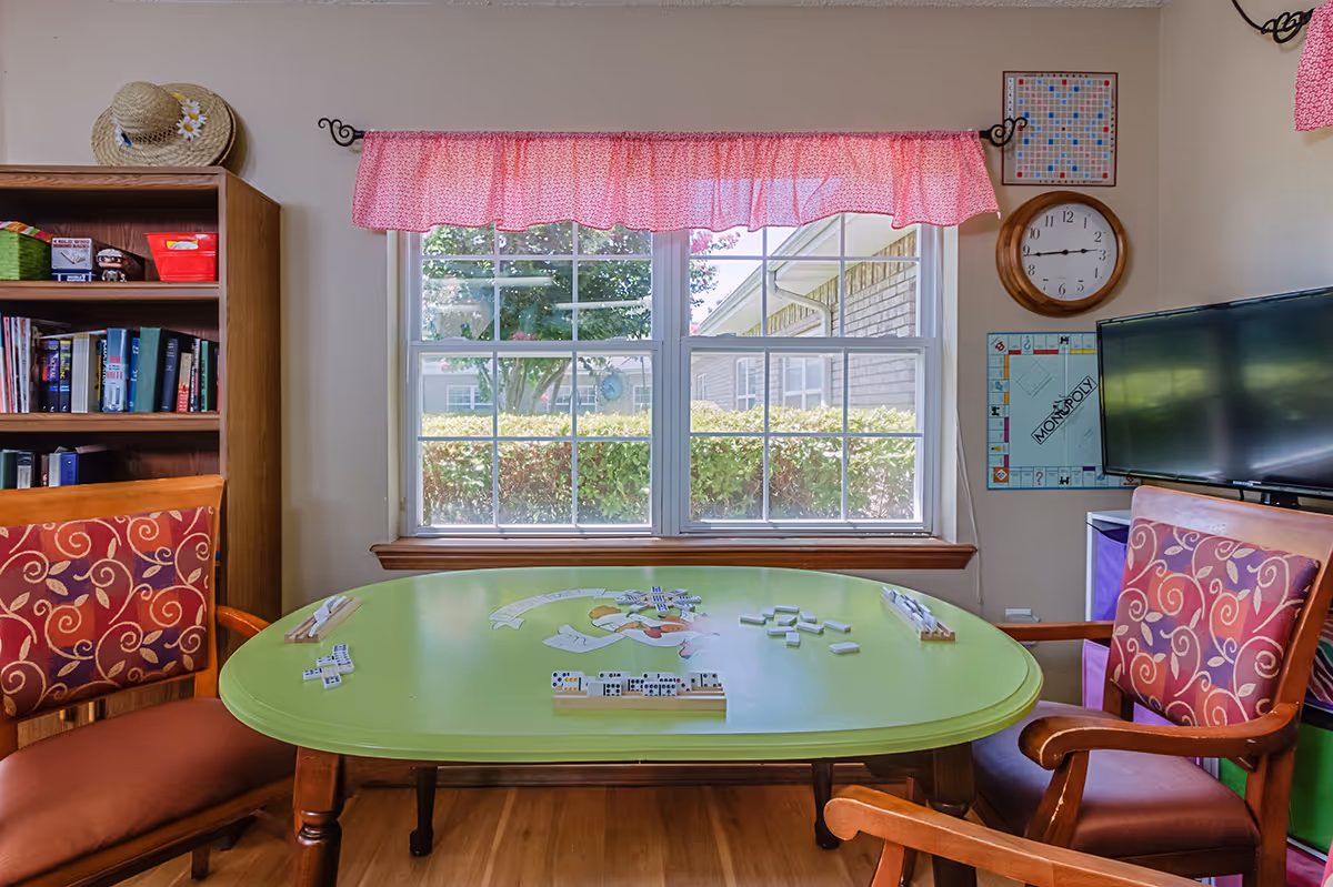 Bright activity room with a green game table set for dominoes, patterned chairs, bookshelf, TV, and a window with a pink valance.