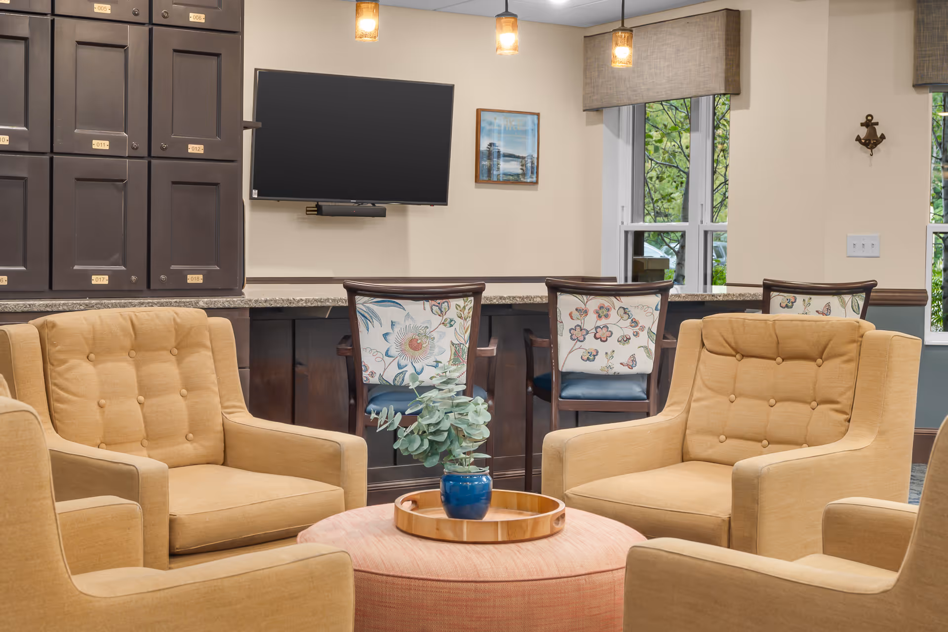 Comfortable common lounge with beige armchairs around a round ottoman in front of a counter with floral-back chairs and a wall-mounted TV.