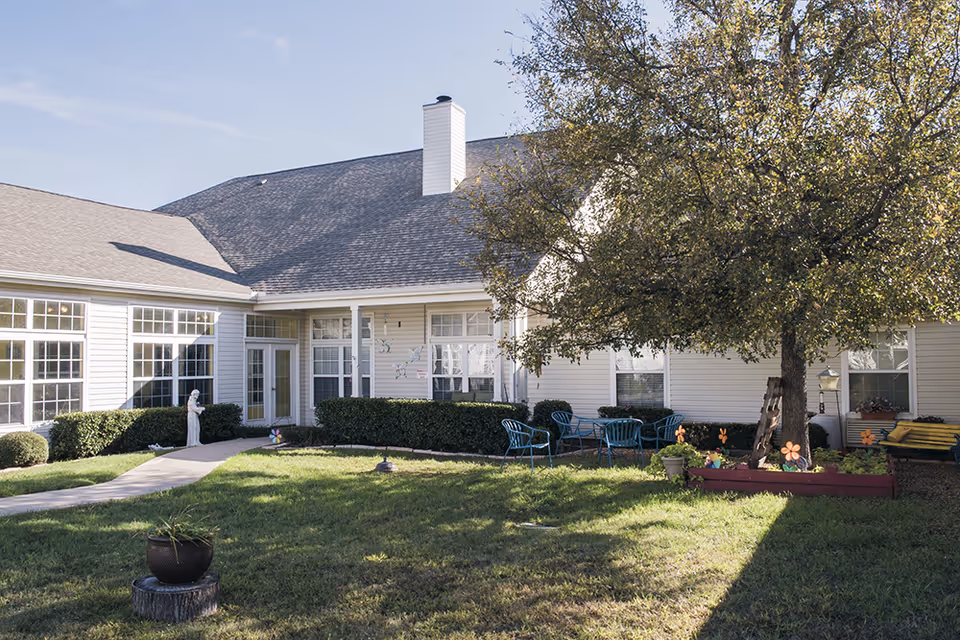 Outdoor courtyard area of a senior living facility with a large tree, patio chairs and tables, a small garden bed with flowers, and a statue near the building entrance. The building is light-colored with multiple windows and a chimney.
